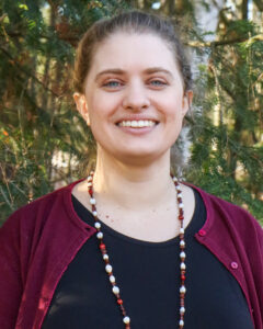 Amanda Strapple, a white female with her hair up in a bun, smiles in front of a sun-dappled evergreen forest while wearing a maroon cardigan over a black top with a long necklace made of brown apple seeds and red and white beads.